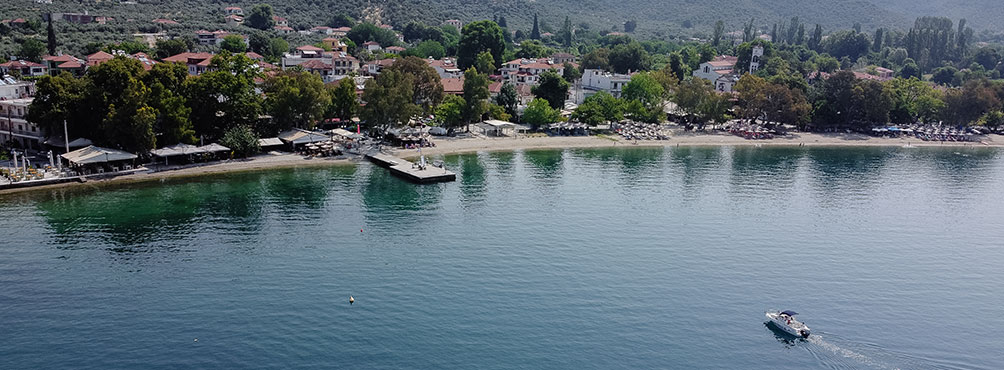 Παραθαλάσσια άποψη του χωριού Άφησσος στο Πήλιο, με αμμώδη παραλία, ταβερνάκια στην ακτή και ένα μικρό σκάφος στη γαλήνια θάλασσα.