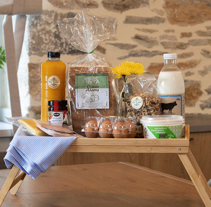 A tray with Greek breakfast essentials, including bread, eggs, milk, granola, orange juice, and local products, showcased by Pelion-villas.com.