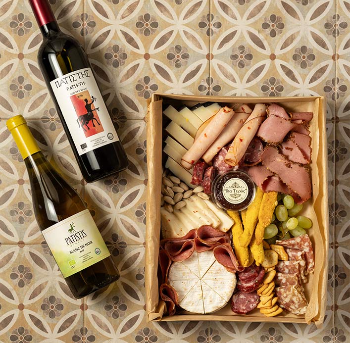 Charcuterie board with assorted Greek cheeses, cured meats, and two bottles of wine on decorative tiles, styled by Pelion-villas.com.