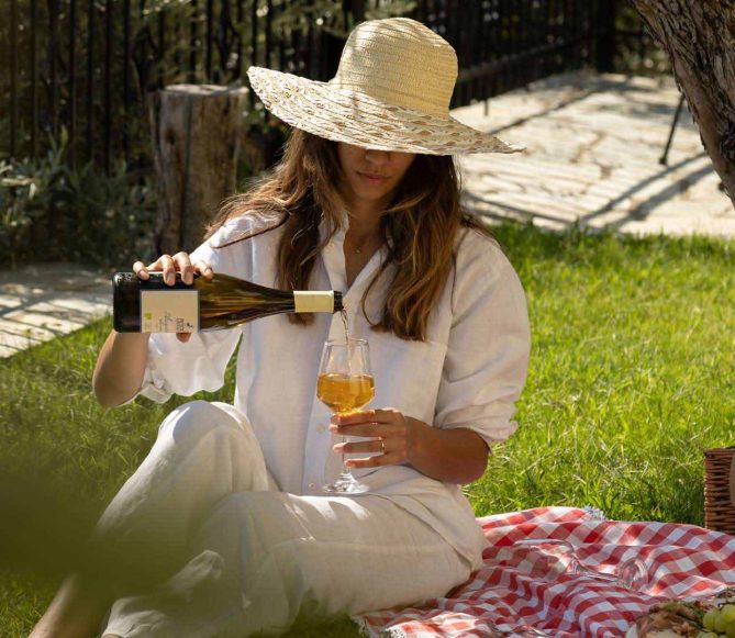 wine-picnic-outdoor-pelion-villas Γυναίκα απολαμβάνει χαλαρωτικό πικνίκ σε γκαζόν με κρασί, ψάθινο καλάθι και ελληνικά σνακ, φιλοξενία από το Pelion-villas.com.
