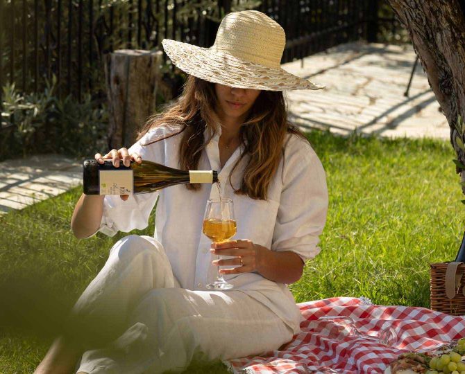 Γυναίκα απολαμβάνει χαλαρωτικό πικνίκ σε γκαζόν με κρασί, ψάθινο καλάθι και ελληνικά σνακ, φιλοξενία από το Pelion-villas.com.