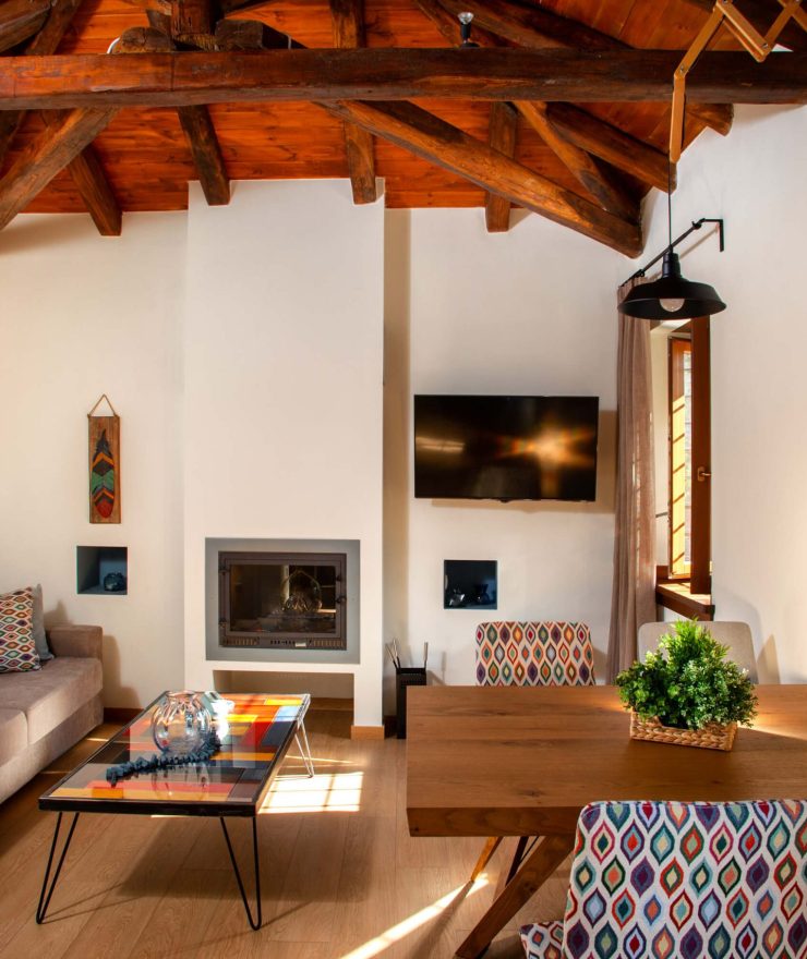 Ένας μοντέρνος και άνετος χώρος καθιστικού και τραπεζαρίας στη Villa Ioanna, με ζεστό τζάκι, πολύχρωμα καθίσματα και ξύλινη οροφή.