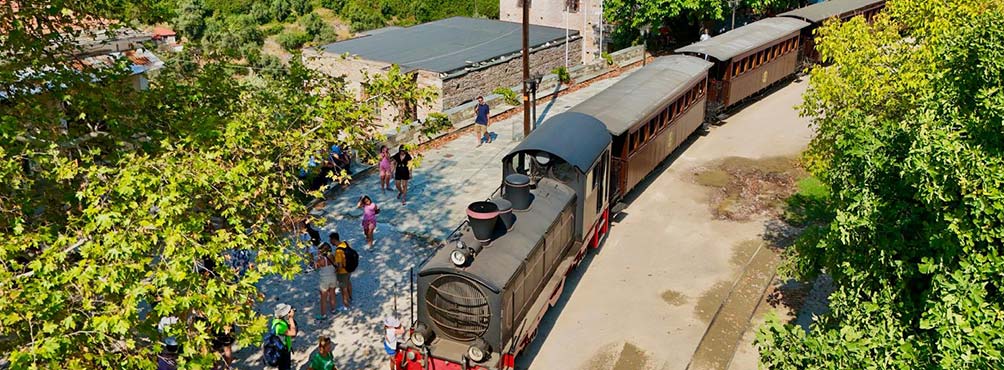 Το ιστορικό τρενάκι του Μουτζούρη στον σταθμό των Μηλεών Πηλίου, με πράσινο περιβάλλον και επισκέπτες έτοιμους για την διαδρομή.