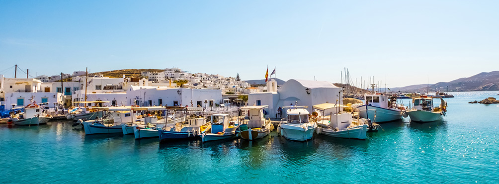 Παραδοσιακά ψαροκάικα σε τιρκουάζ νερά μπροστά από λευκά κτίρια στο λιμάνι της Νάουσας στην Πάρο.