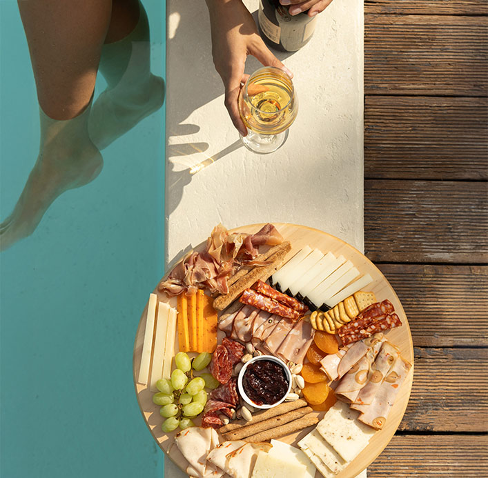 Poolside cheese and charcuterie board with cured meats, Greek cheeses, fresh fruits, and a glass of white wine, captured at Pelion-villas.com.