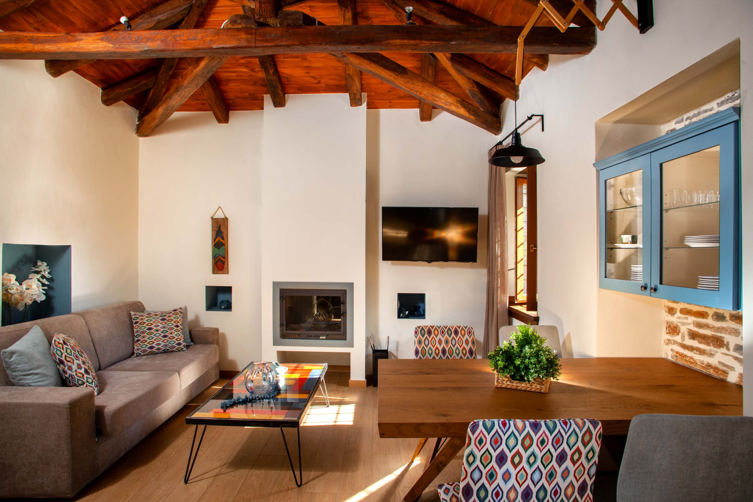 A stylish and cozy living and dining area at Villa Ioanna, featuring a warm fireplace, colorful seating, and a wooden ceiling.